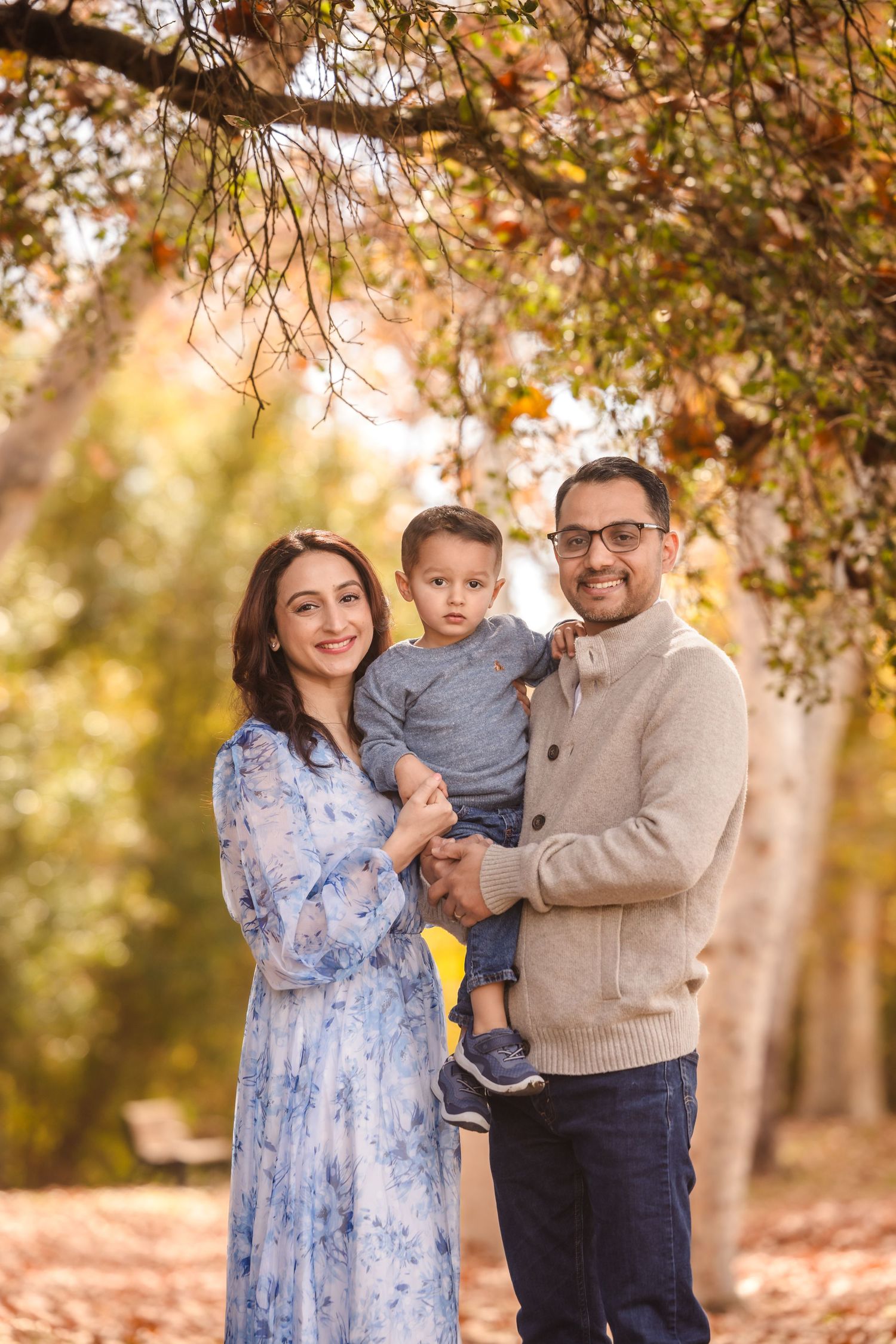 A family of three smiles warmly during their fall photoshoot in a wooded park.