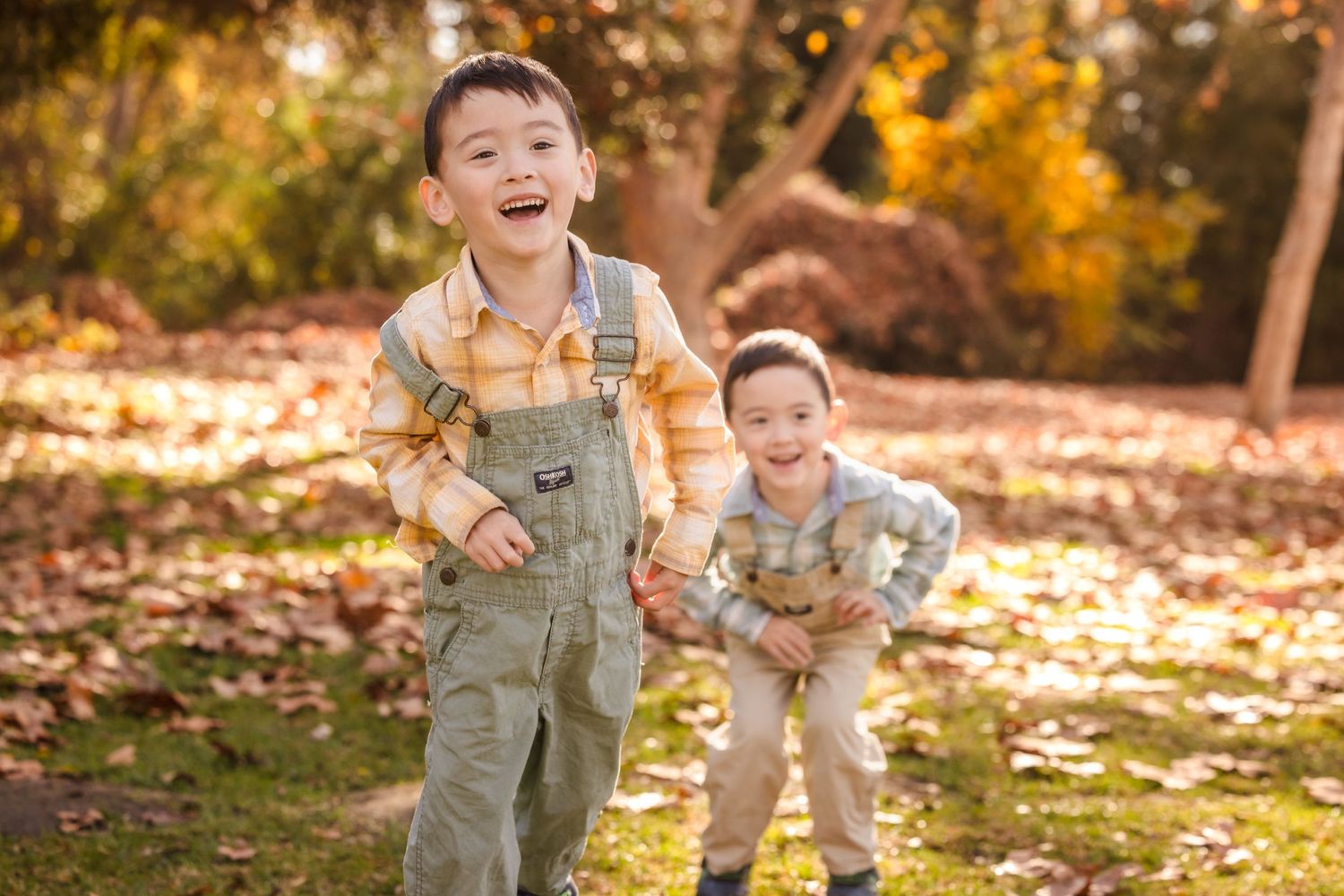 Two children in overalls play and laugh in autumn setting with golden sunlight.