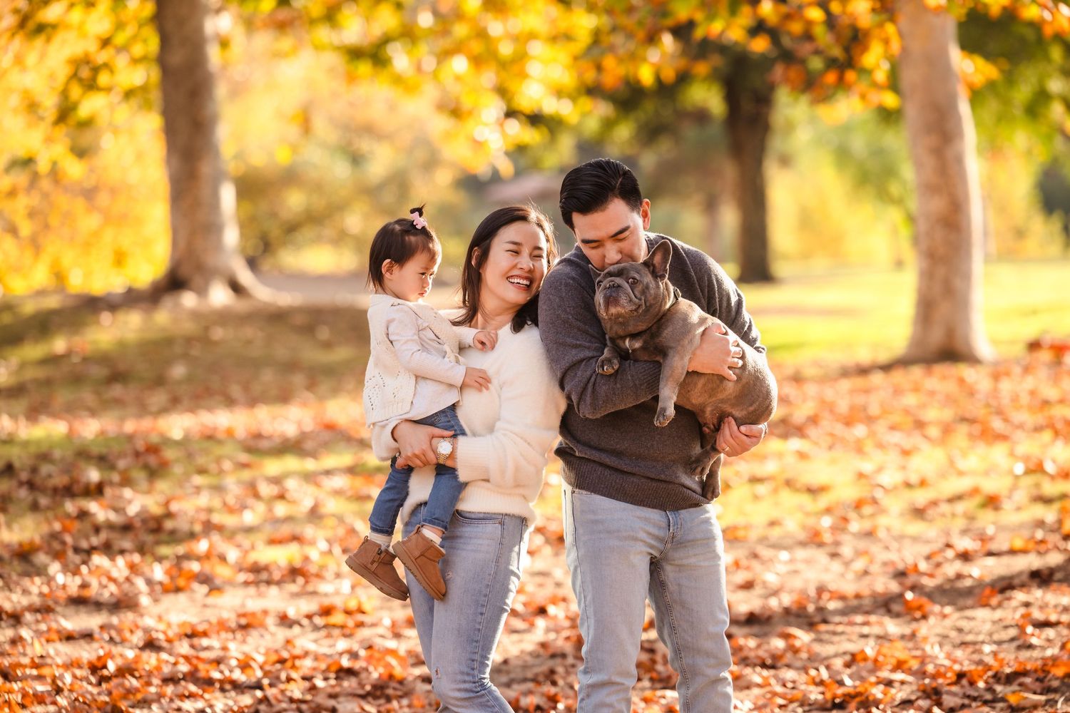 A family walks together with their small brown dog through a park filled with golden autumn leaves.