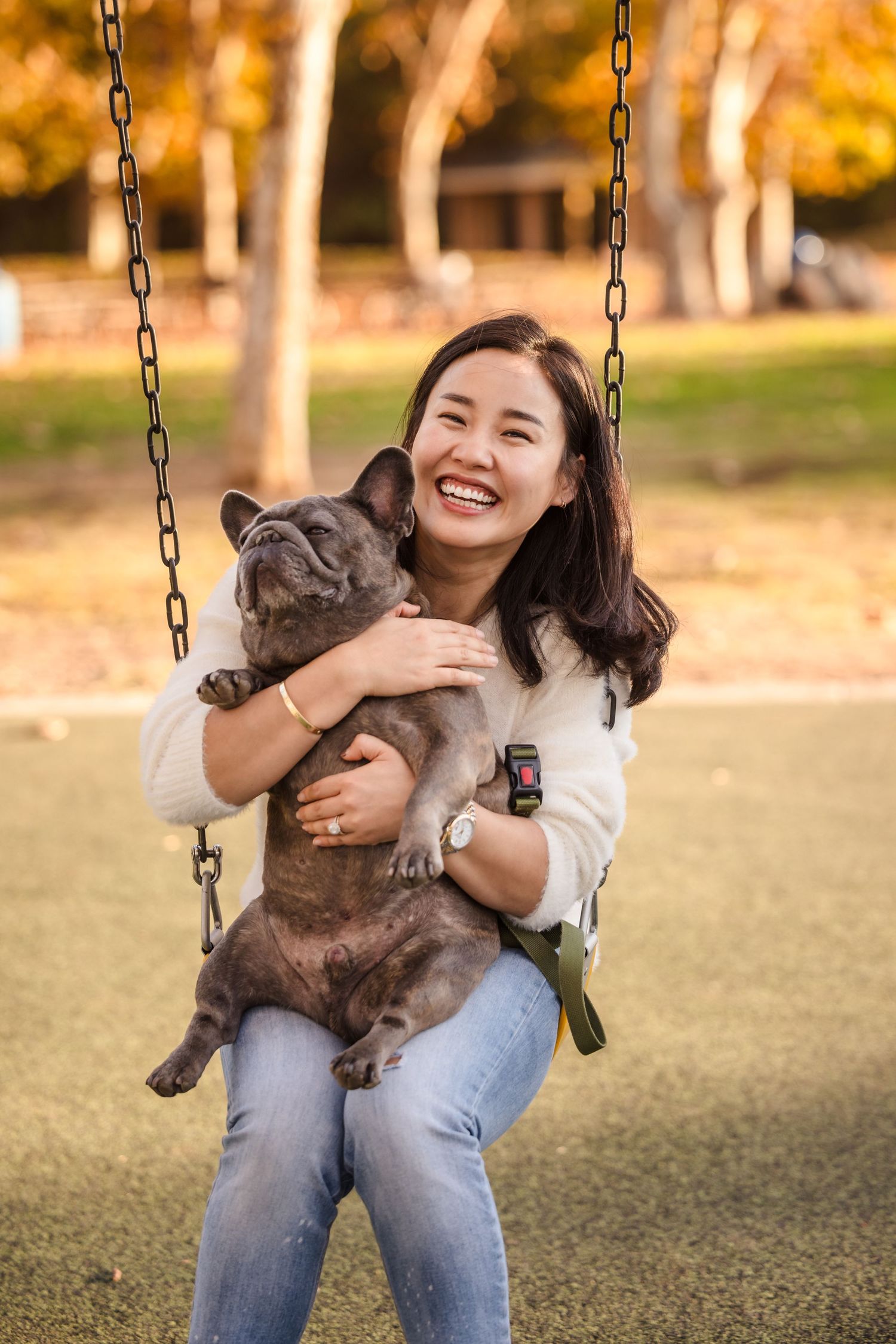A person sits on a swing holding a French Bulldog in a park with autumn trees.