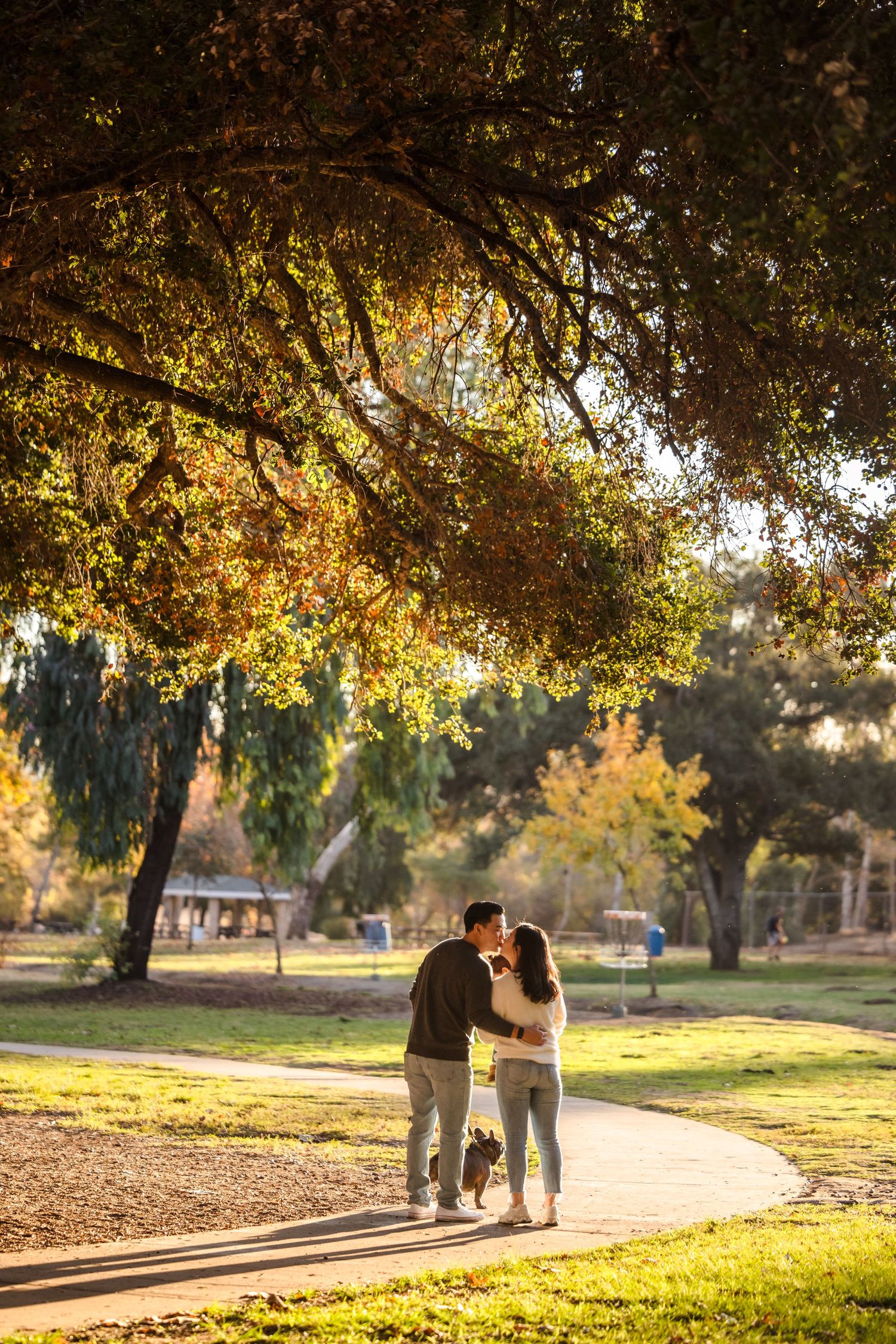 A couple walks together on a park path under golden autumn trees during sunset.
