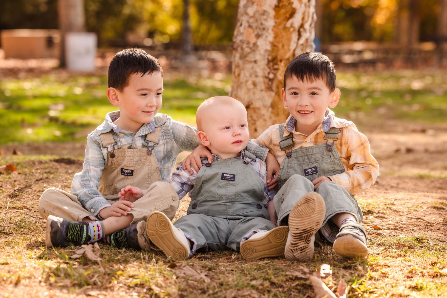 Three young children sit together on fallen leaves during autumn photoshoot.