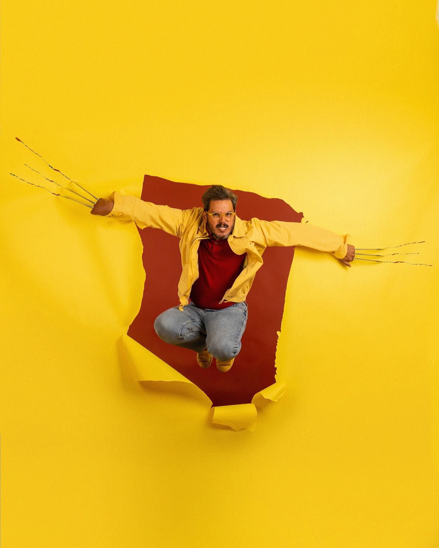 A person bursts through yellow paper wall wearing red sweater and jeans while striking a dynamic jumping pose.