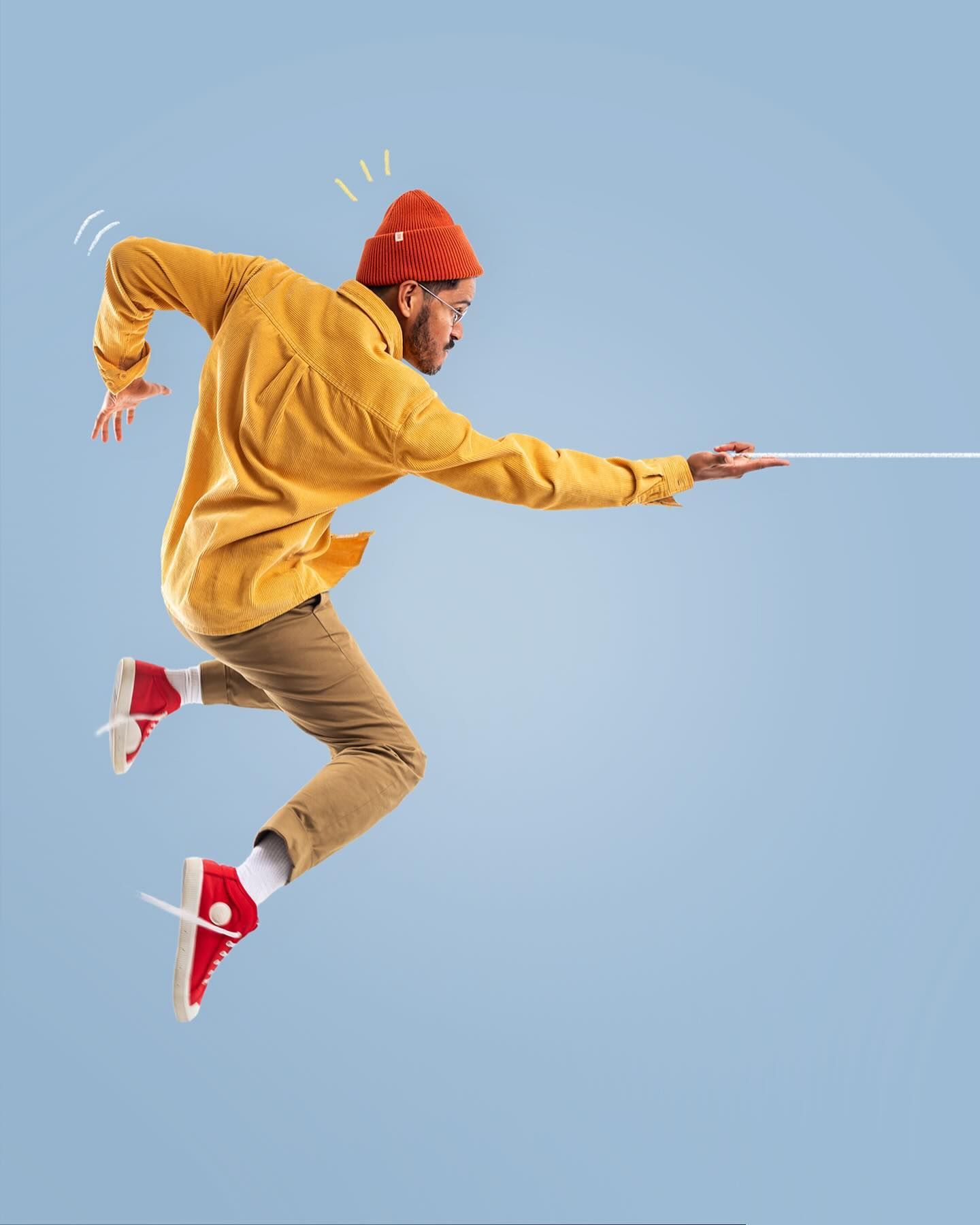 Person in yellow hoodie and red beanie jumping sideways against blue background while pulling invisible rope.