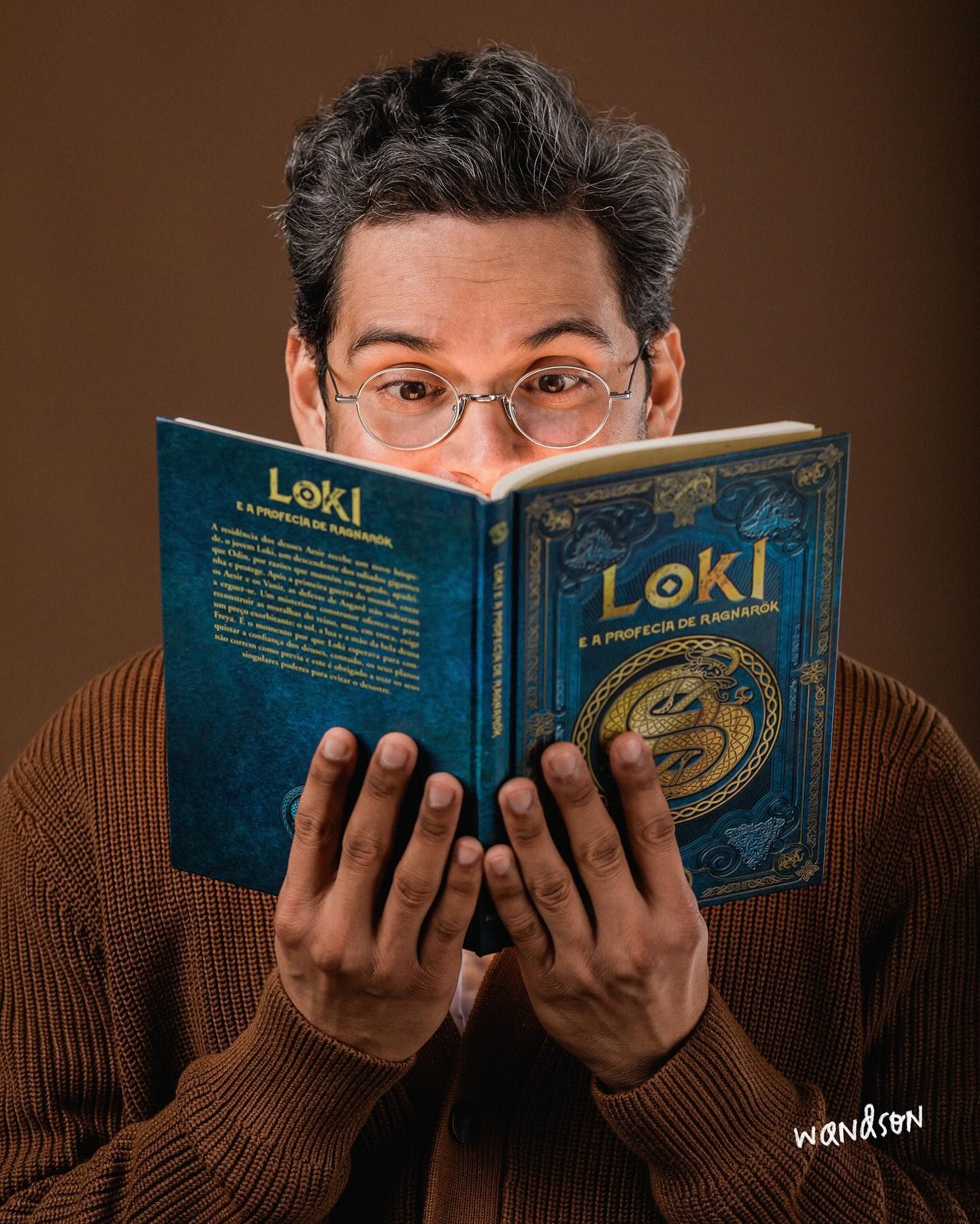 Someone in a brown sweater holds up and reads from a hardcover book titled 'Loki' with a blue and gold cover design.