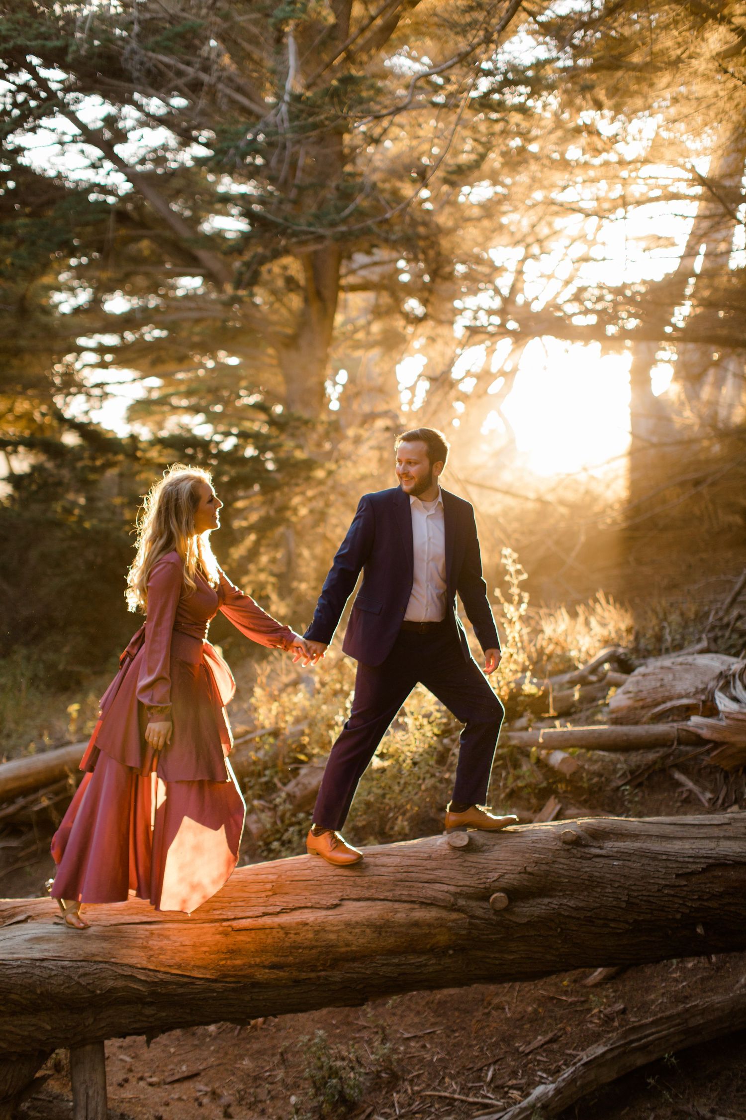 A couple balances on a fallen log in a sunlit forest during golden hour.