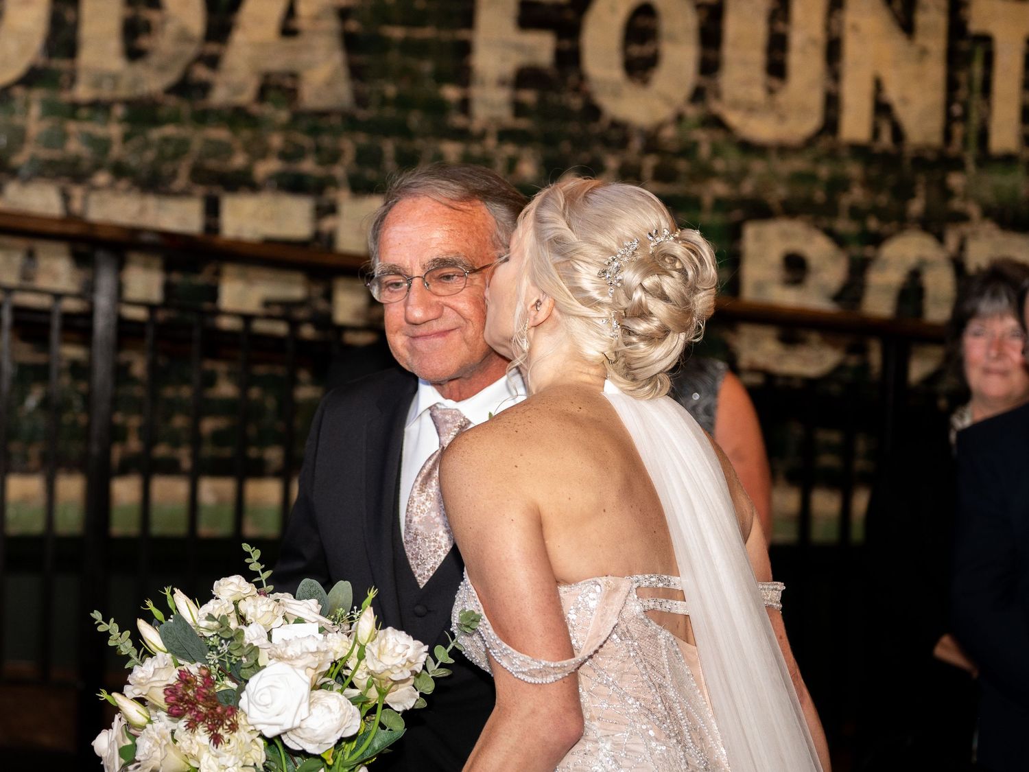 A bride in a strapless gown with a bouquet smiles beside someone at an outdoor venue with text visible in background.