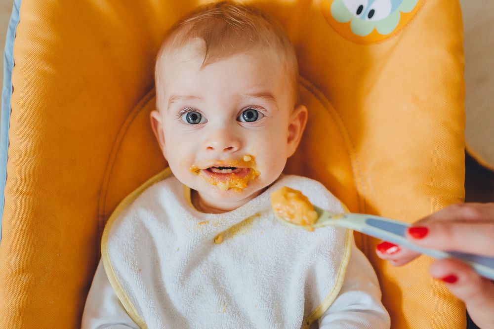 Baby in einem Hochstuhl, trägt Weiß und hat Essen um den Mund, während es mit einem rot-griffigen Löffel gefüttert wird.