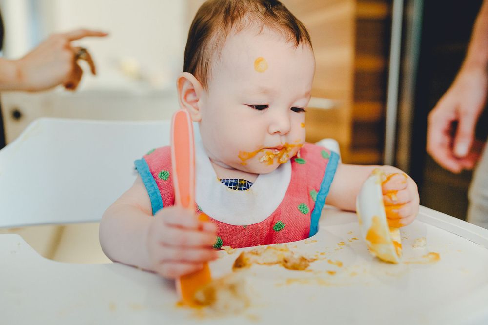 Baby im Lätzchen sitzt im Hochstuhl und spielt mit Essen, während es Selbstfüttern während der Mahlzeit lernt.