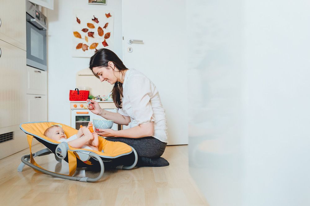 Elternteil sitzt auf dem Boden und interagiert mit dem Baby in einem Schaukelstuhl in einem hell erleuchteten, minimalistischen Raum.