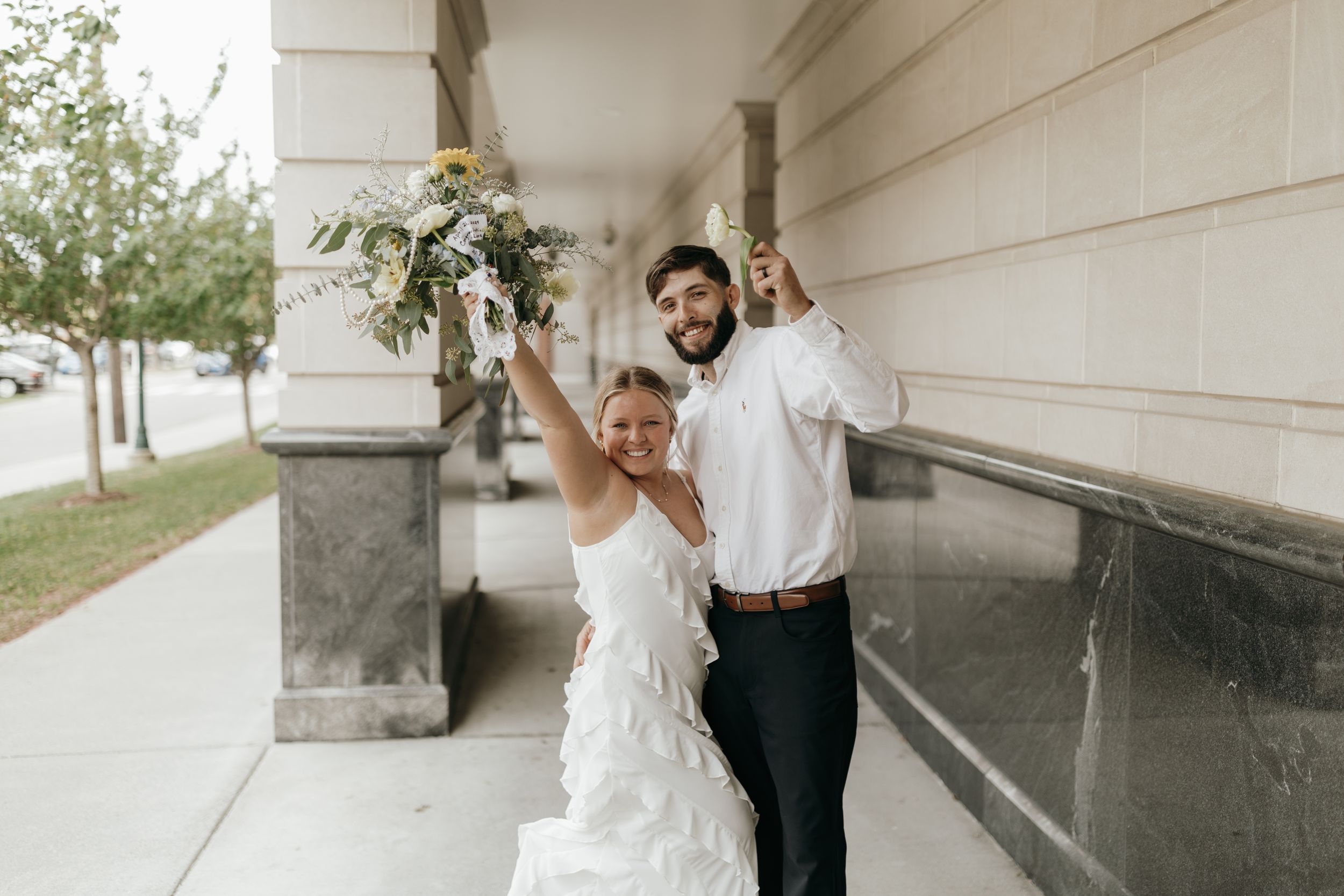Joyful wedding couple celebrating with raised arms against a stone column.
