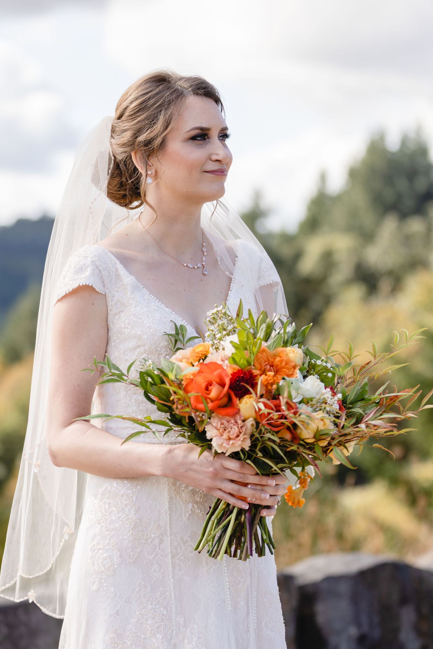 A bride in a lace dress holds a vibrant orange and peach floral bouquet against a mountain backdrop.