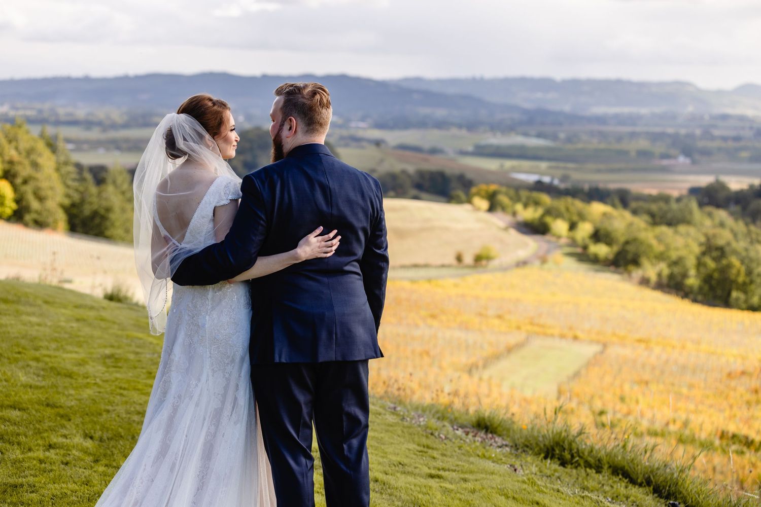 A couple embraces while looking out over rolling hills and golden fields on their wedding day.