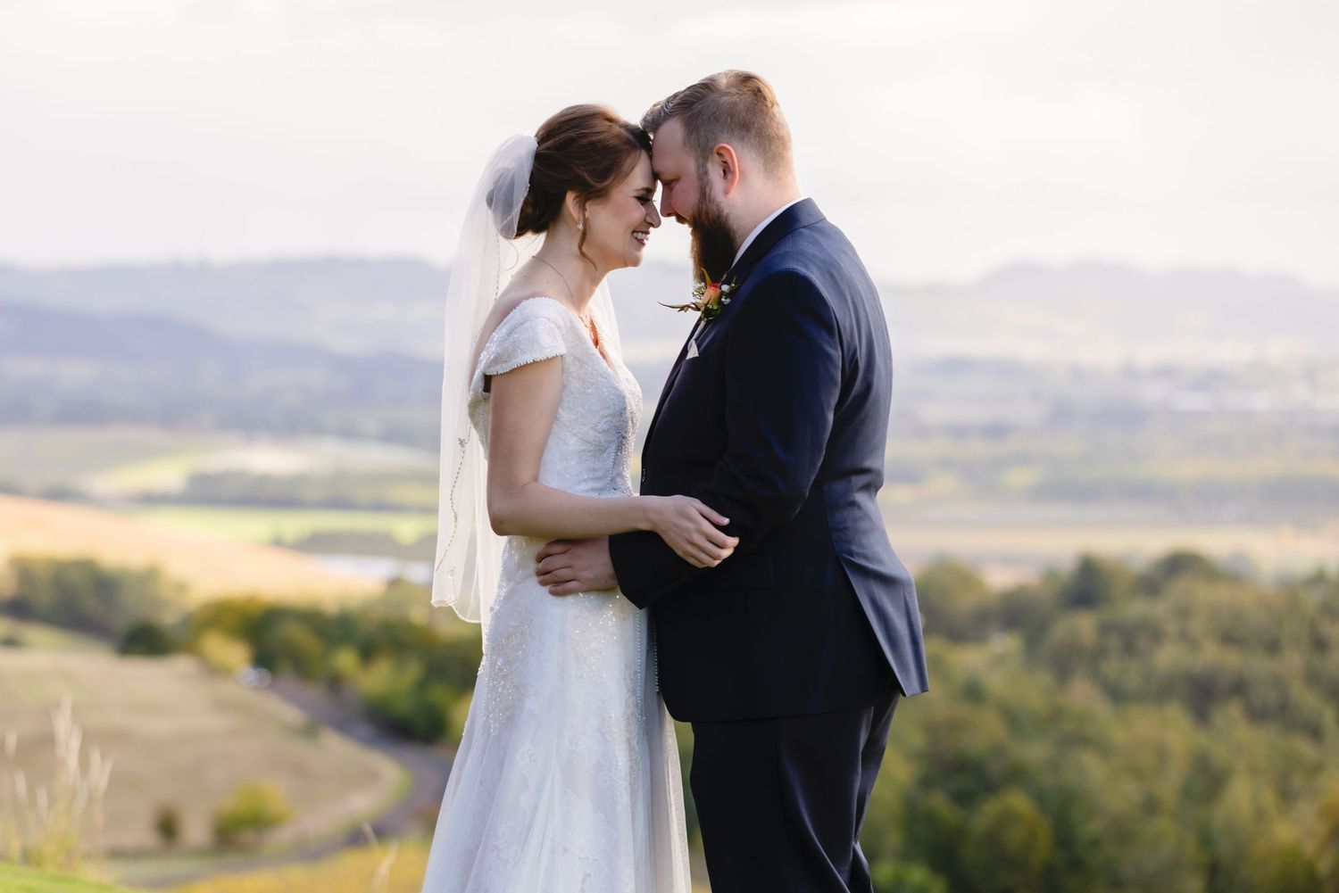 A couple in wedding attire embraces while standing against a scenic mountain landscape at sunset.