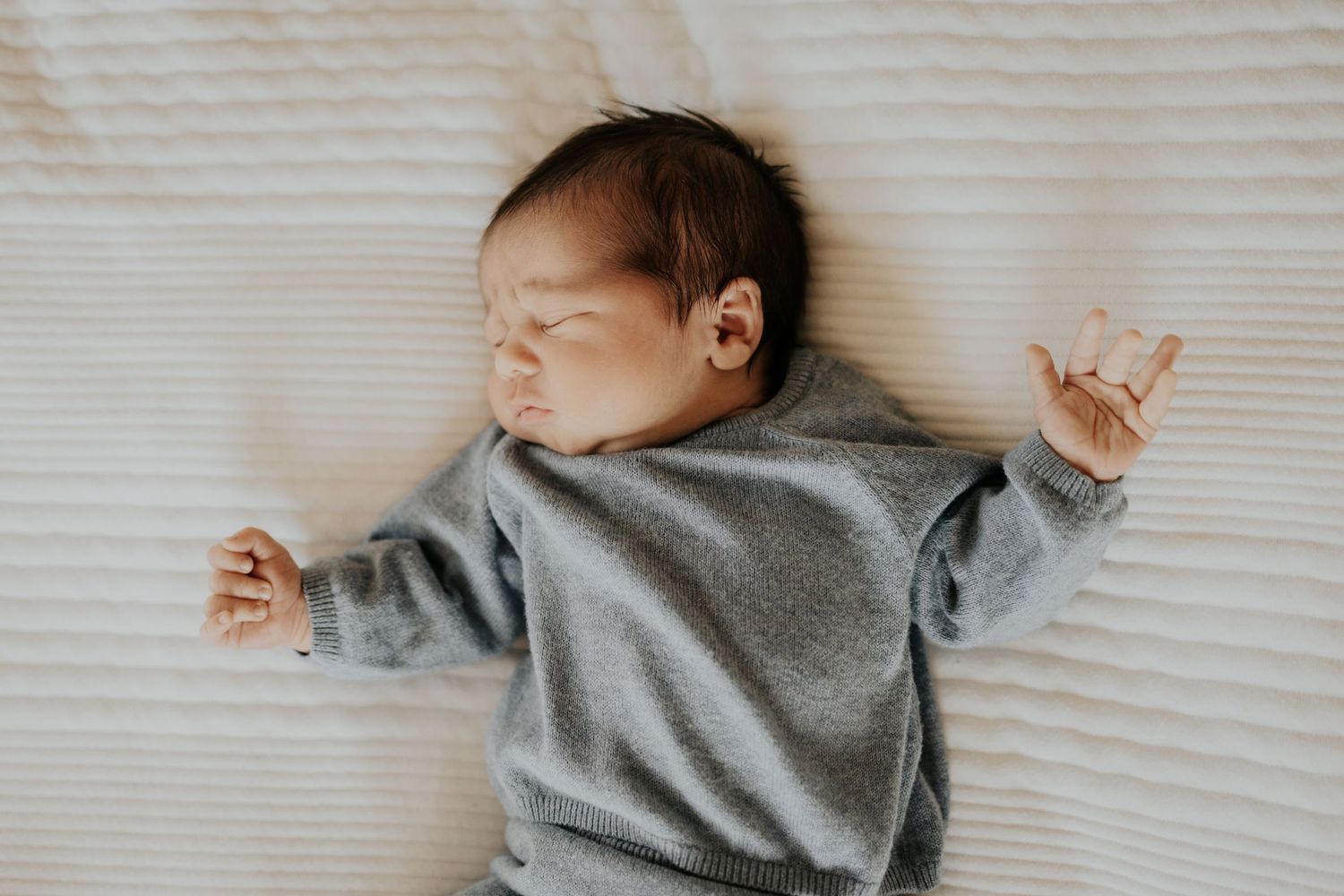 Sleeping newborn baby wearing grey sweatshirt lying on white textured blanket with arms outstretched.