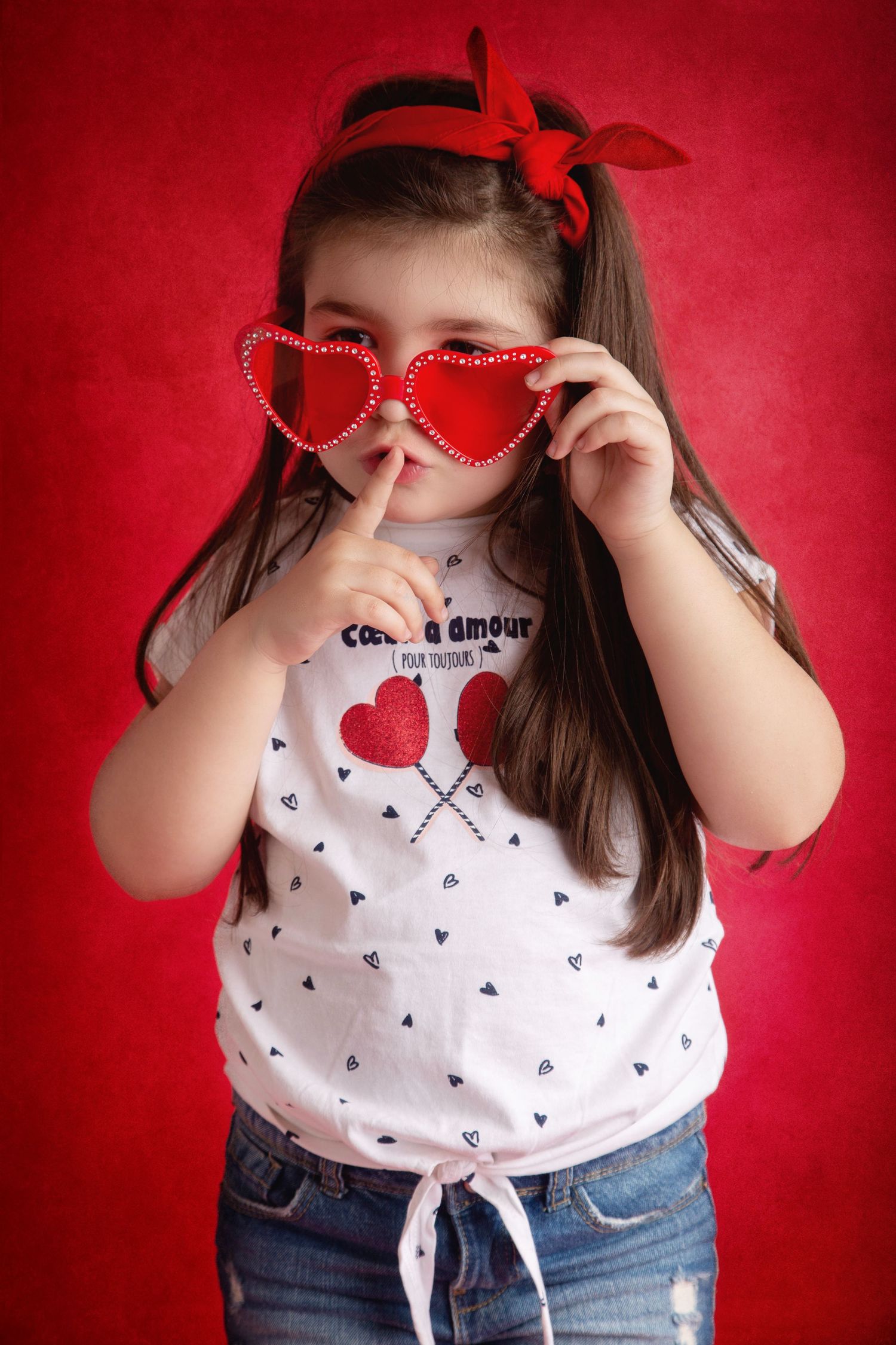Playful Valentine's Day portrait against red background with heart sunglasses and matching bow accessory.