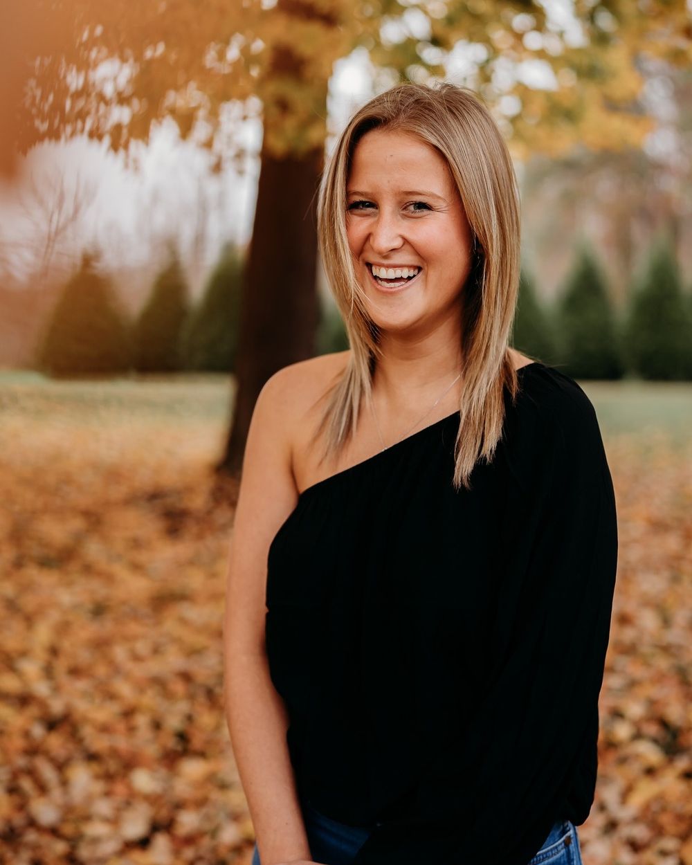 Portrait of woman in black one-shoulder top smiling in autumn setting with fallen leaves.