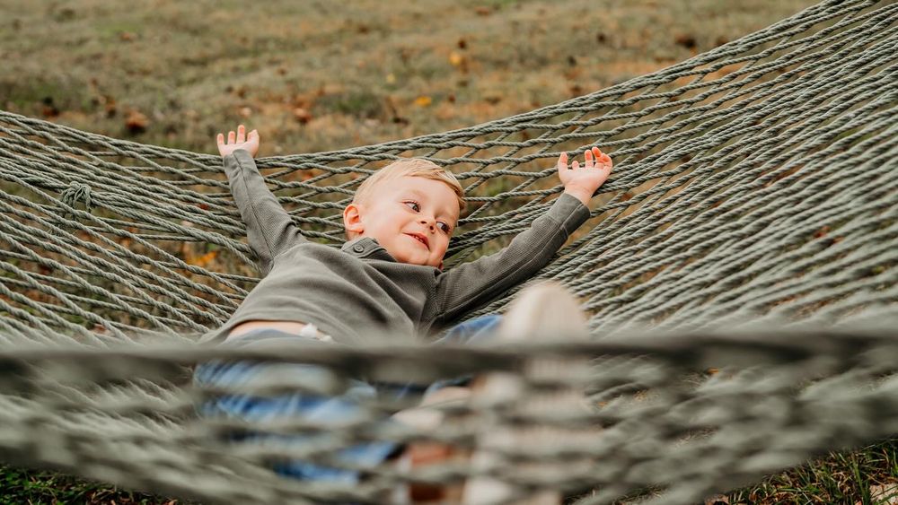 Child lying in hammock with arms raised against autumn background.