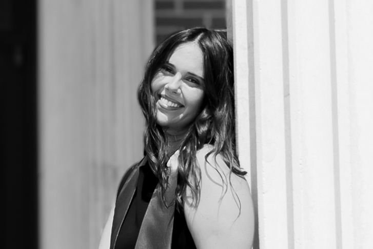Black and white photo of a smiling brunette woman leaning against a white pillar outdoors.