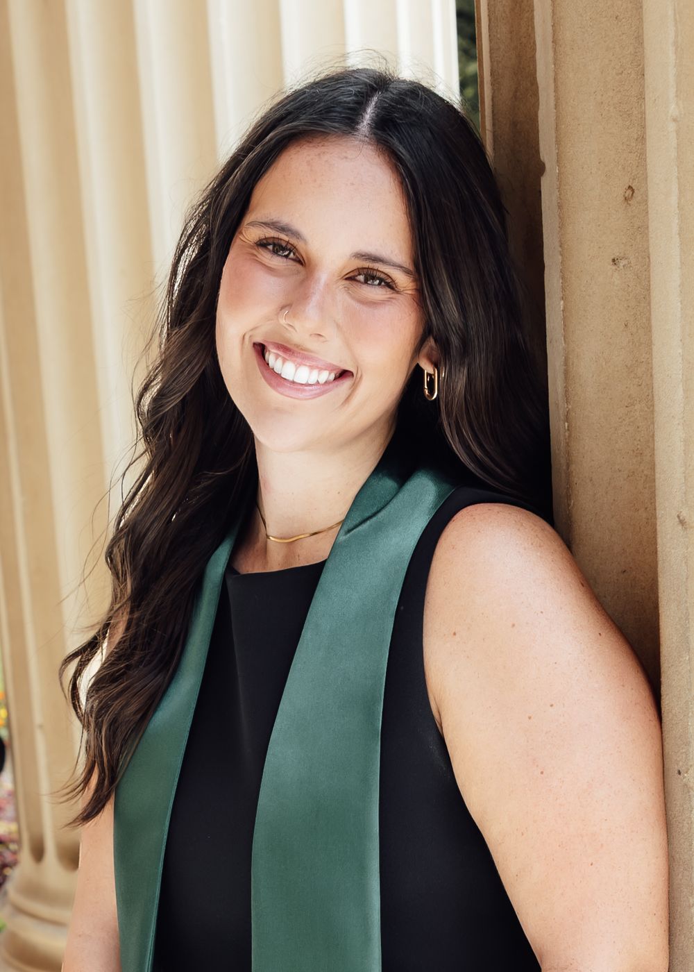 Smiling young woman with long dark hair wearing a black top and green sash, posing beside elegant stone columns outdoors.