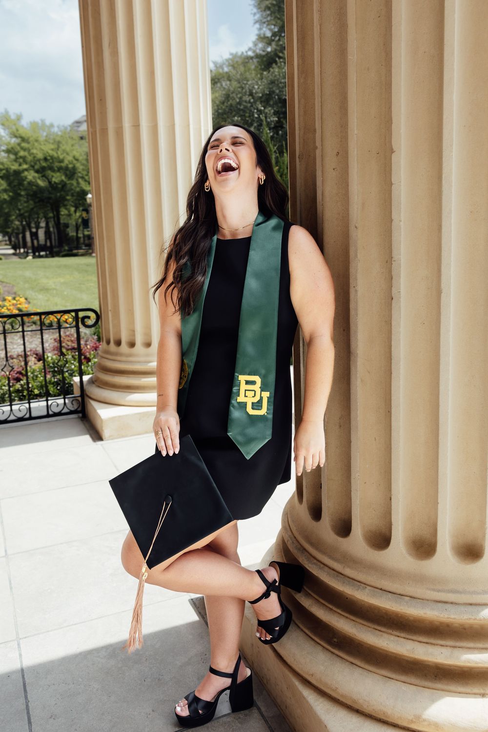 Baylor University graduate laughing joyfully, wearing green BU stole and holding cap, posed by classical columns outdoors.