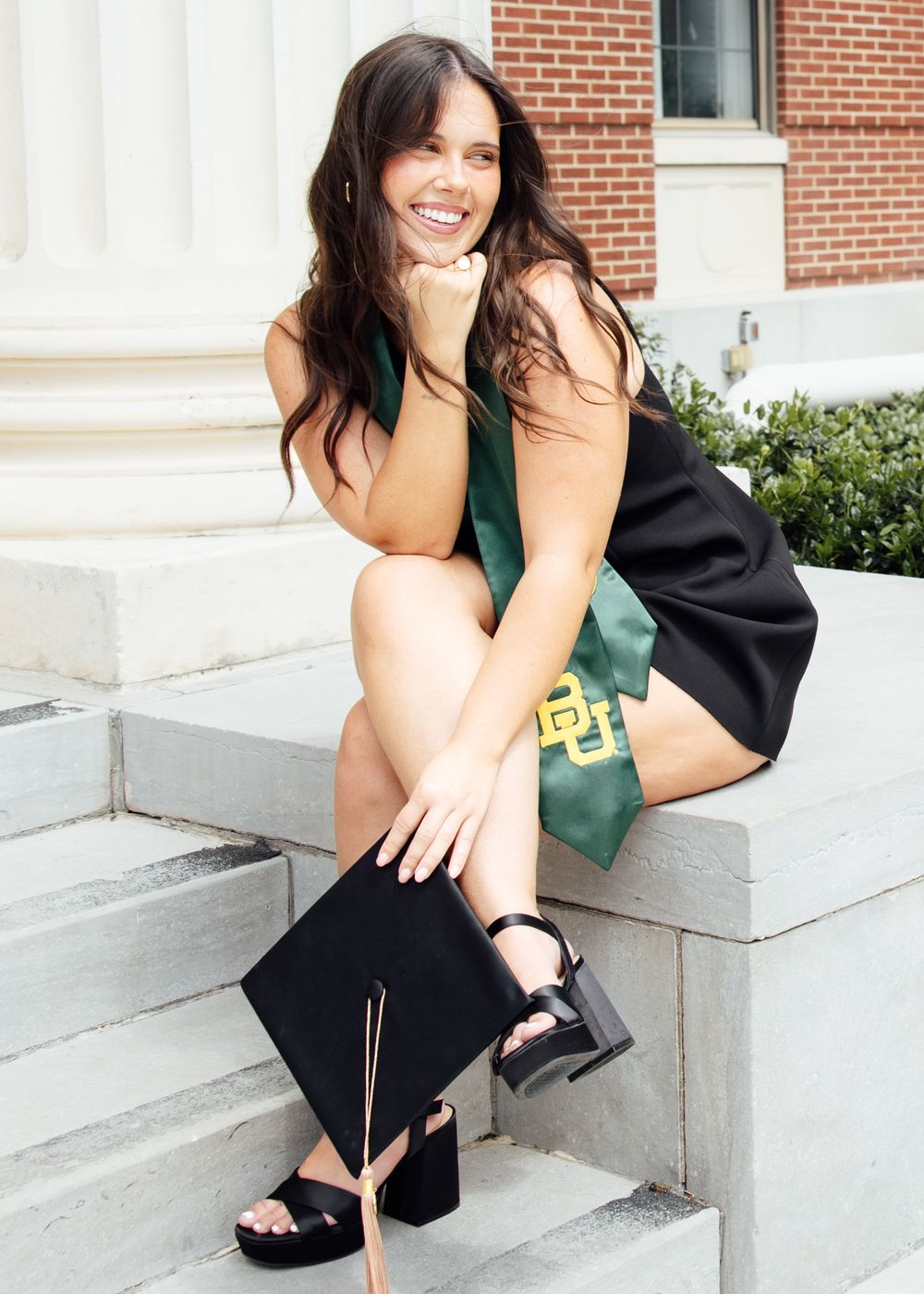 Smiling female graduate sitting on steps wearing black dress and green Baylor University stole holding graduation cap.
