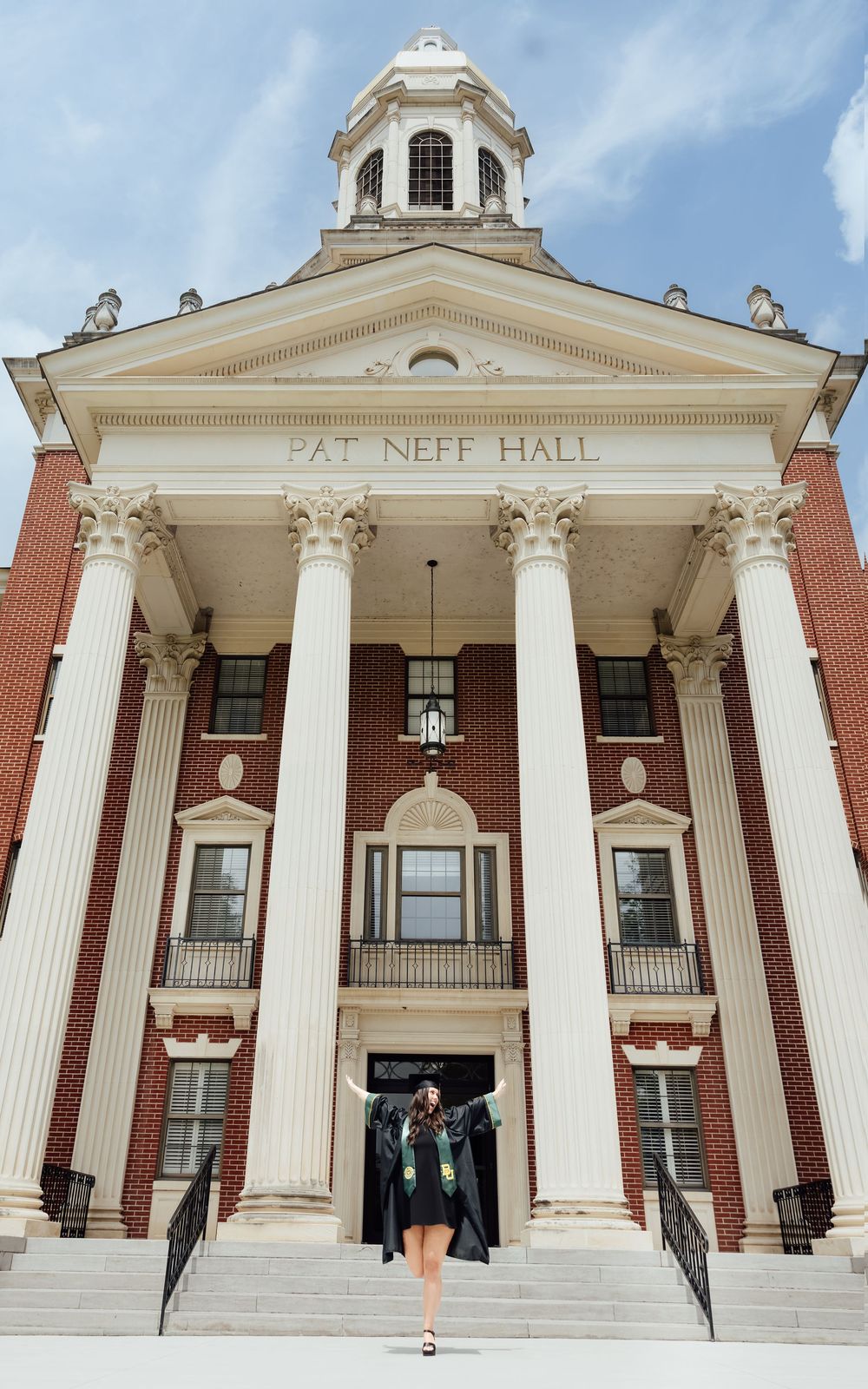 Graduate poses on steps of Pat Neff Hall, a historic brick building with white columns at Baylor University.