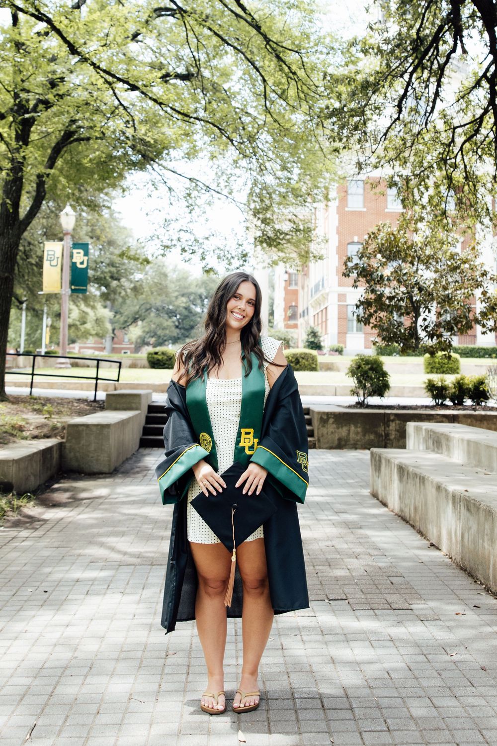 Smiling female graduate wearing BU stole and open gown poses on Baylor University campus walkway.