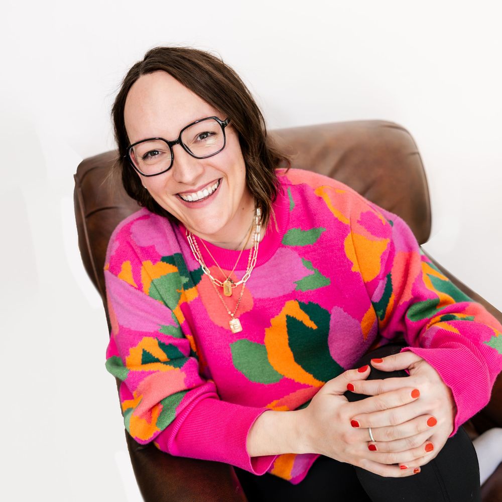 Smiling woman in colorful pink floral sweater with glasses seated in brown leather chair.