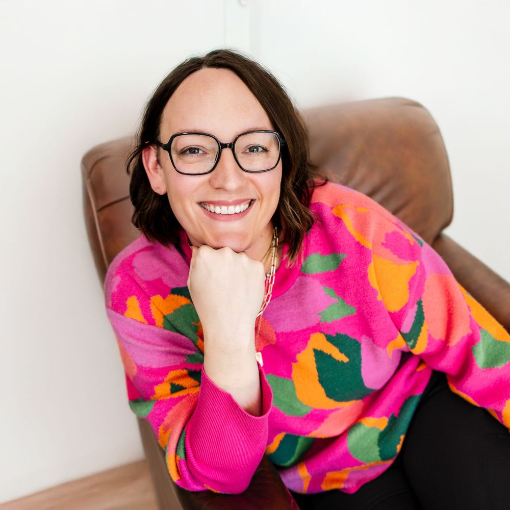 Smiling woman in colorful pink sweater with glasses leaning on brown leather chair.