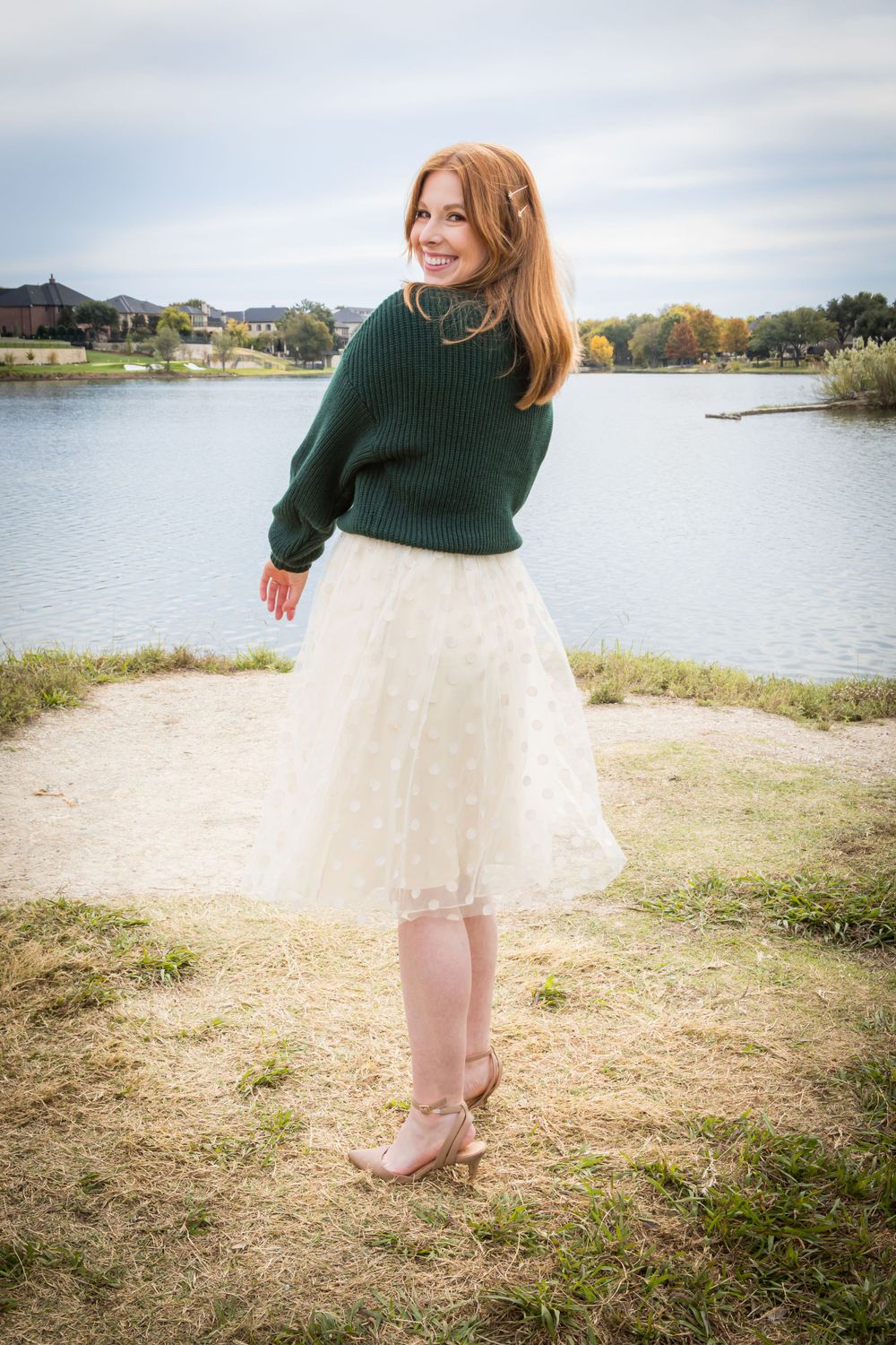Joyful redhead woman in a dark green sweater and ivory tulle skirt smiling by a serene lakeside.