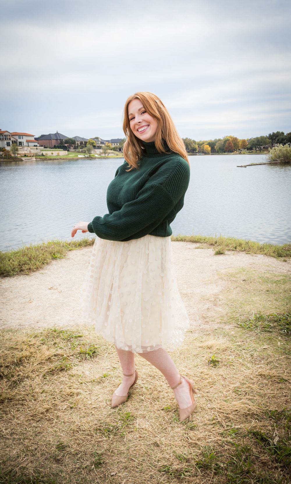 Cheerful redhead woman in a green turtleneck sweater and white lace skirt laughing by a calm lake.