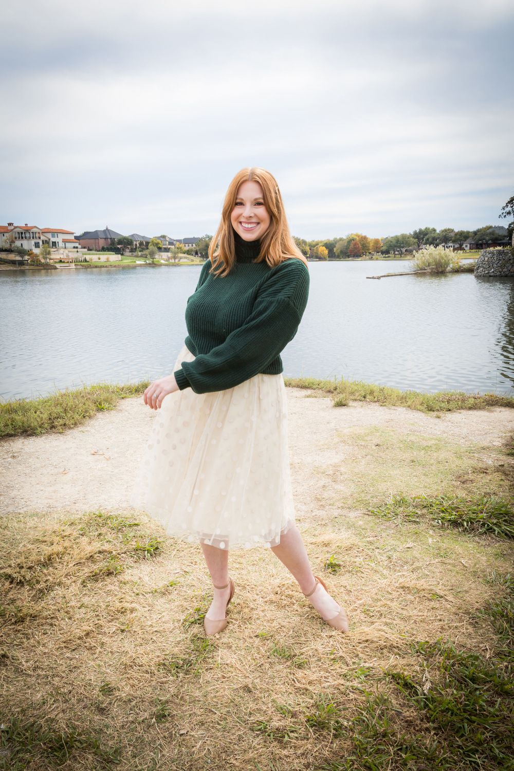 Laughing redhead woman in a green sweater and white tulle skirt posing playfully near a peaceful lake.