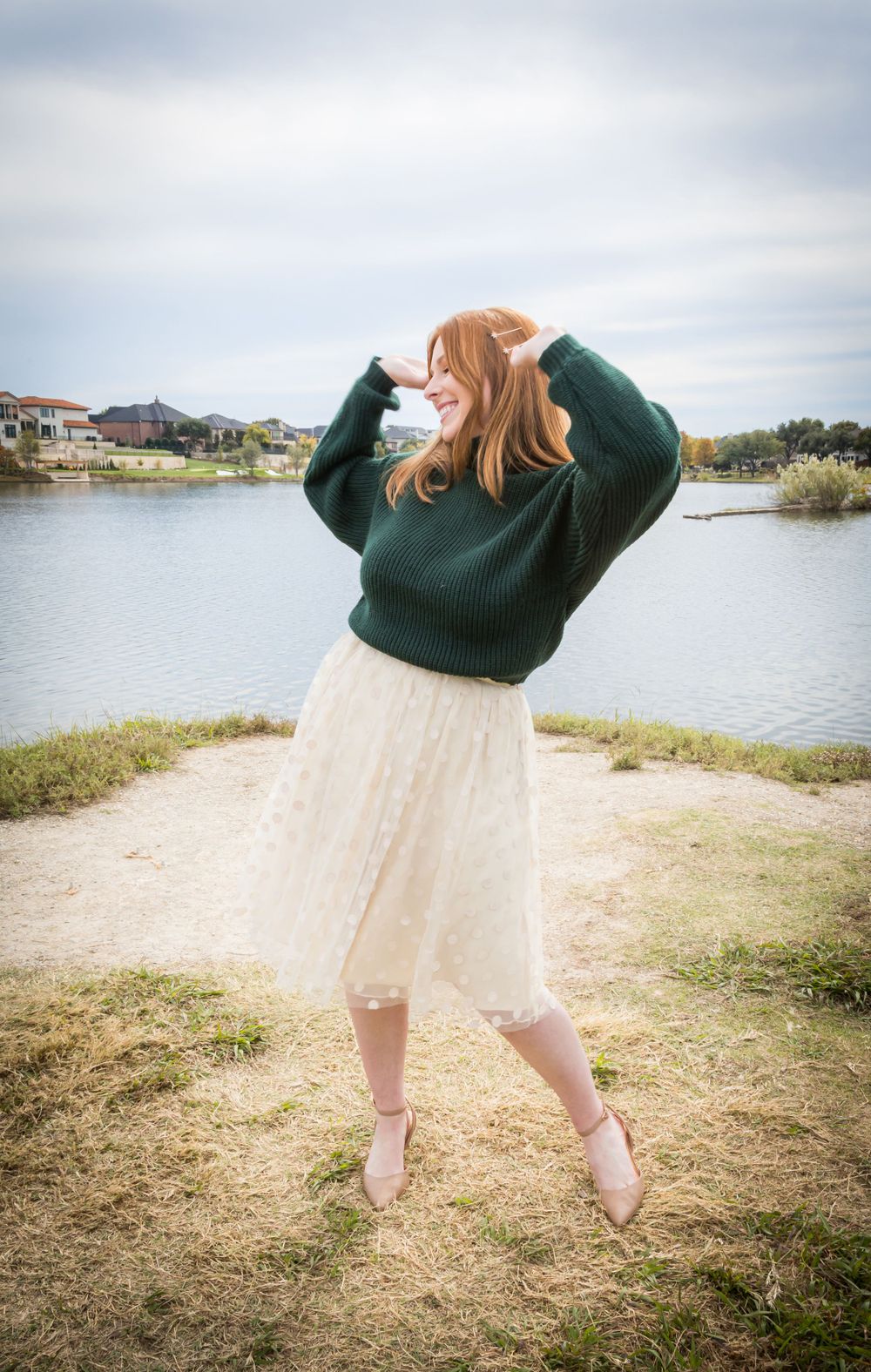 Redhead woman in a green sweater and white midi skirt playfully tosses her hair by a lakeside.