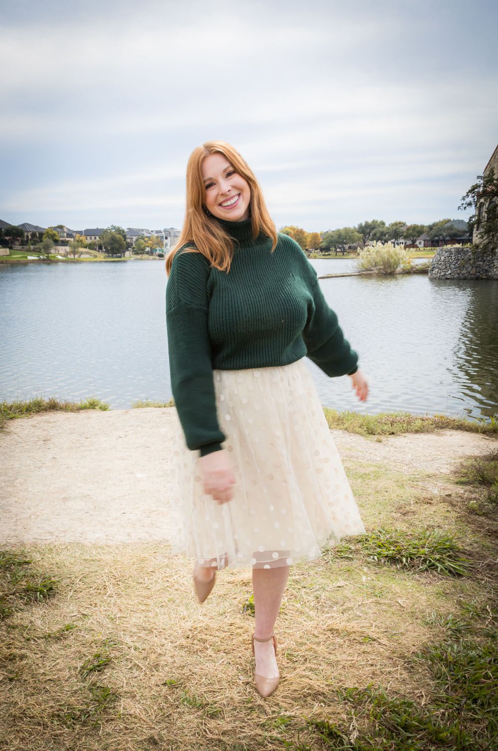 Happy redhead woman in a green sweater and ivory tulle skirt smiling and posing by a scenic lake.