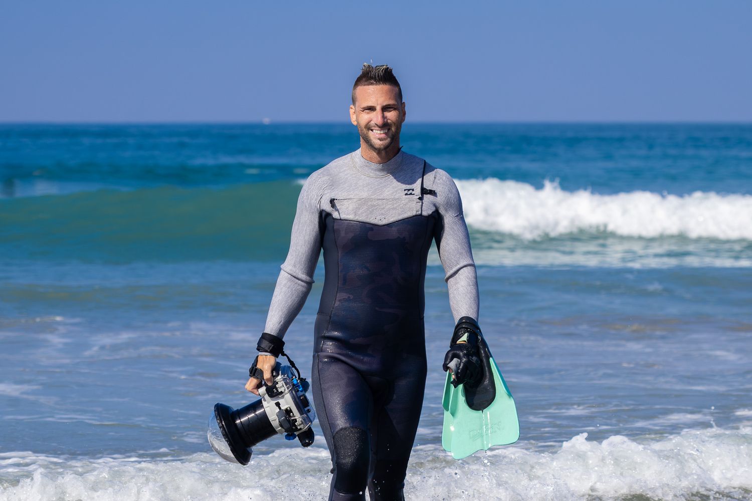 San Diego surf photographer Steve in a wetsuit wading through ocean waves holding swim fins and an underwater camera housing.