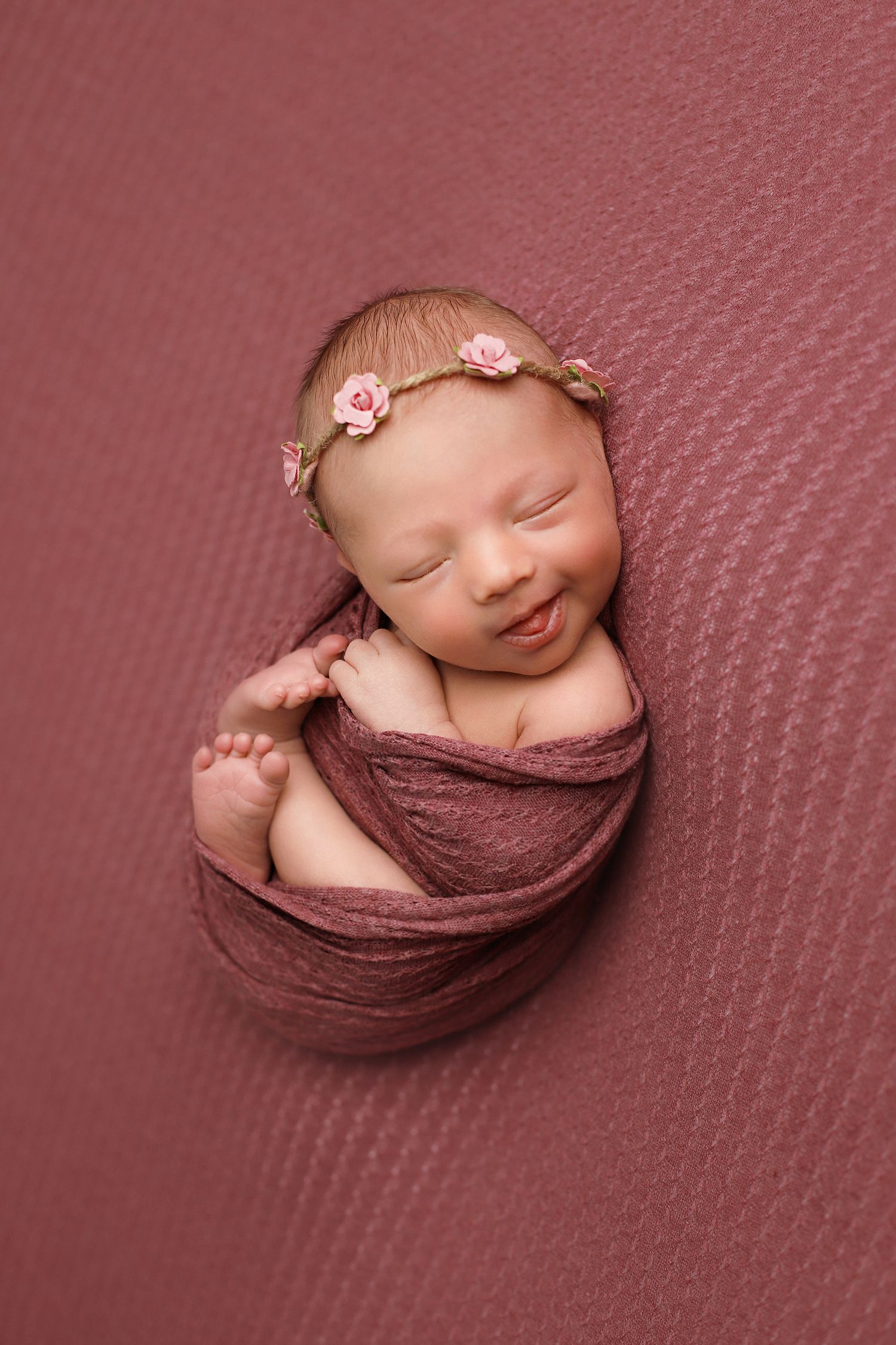 Newborn baby swaddled in mauve fabric wearing delicate pink floral headband sleeps peacefully on textured backdrop.