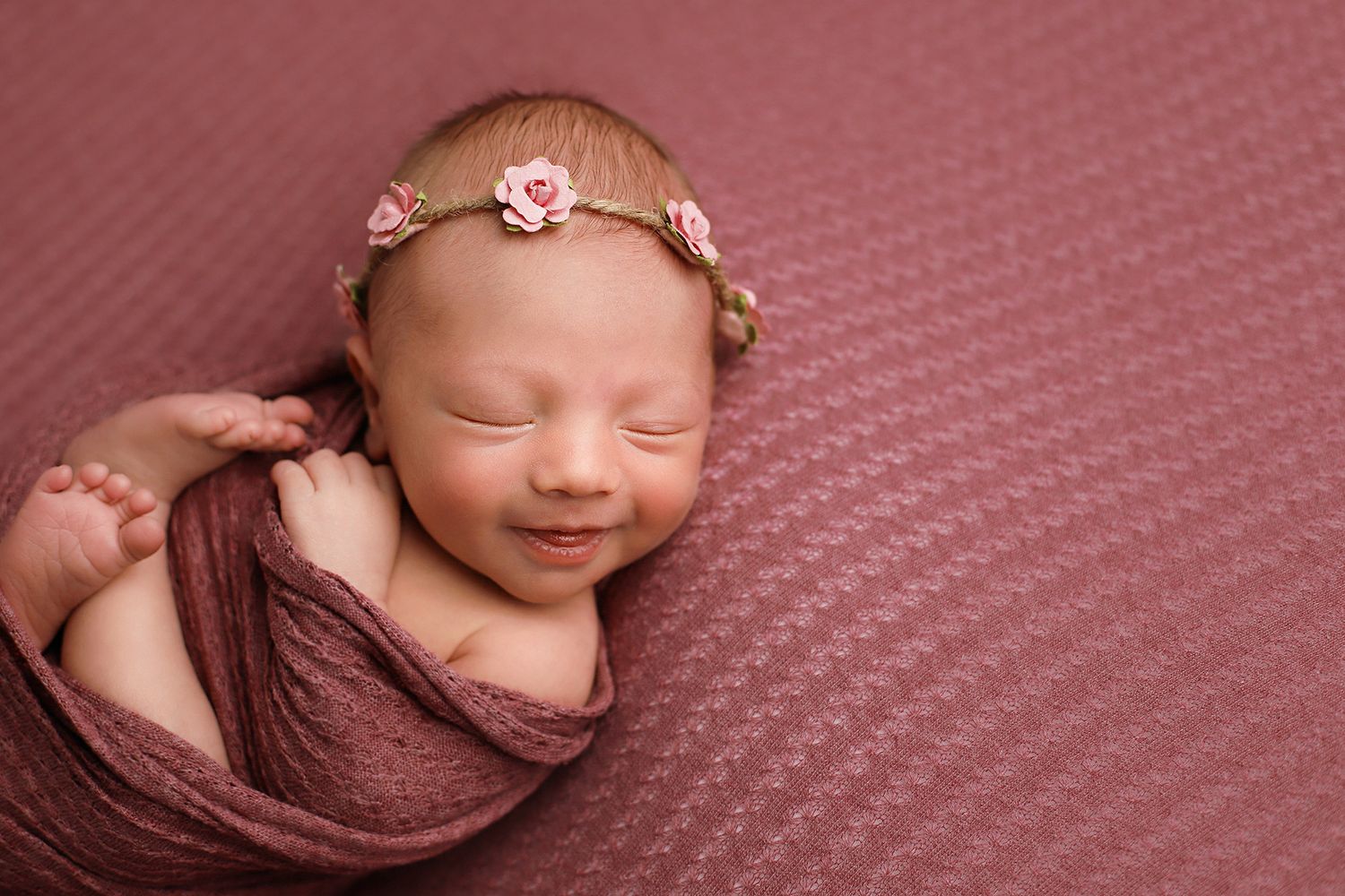 Newborn baby swaddled in brown fabric sleeping peacefully with tiny floral headband on pink textured backdrop.