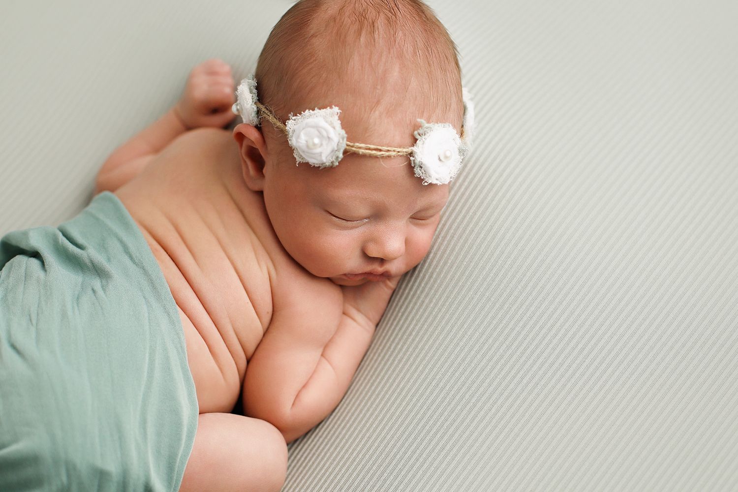 Sleeping newborn wearing delicate white floral headband wrapped in mint green fabric on textured gray background.