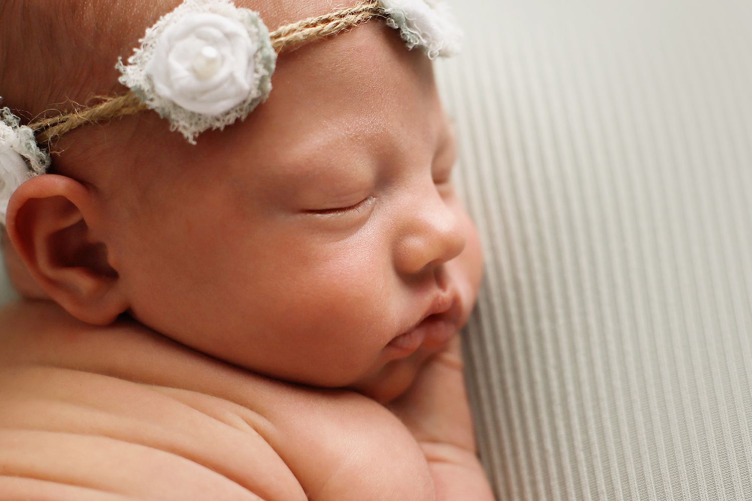 Peaceful sleeping newborn wearing delicate white floral headband resting on soft textured fabric.