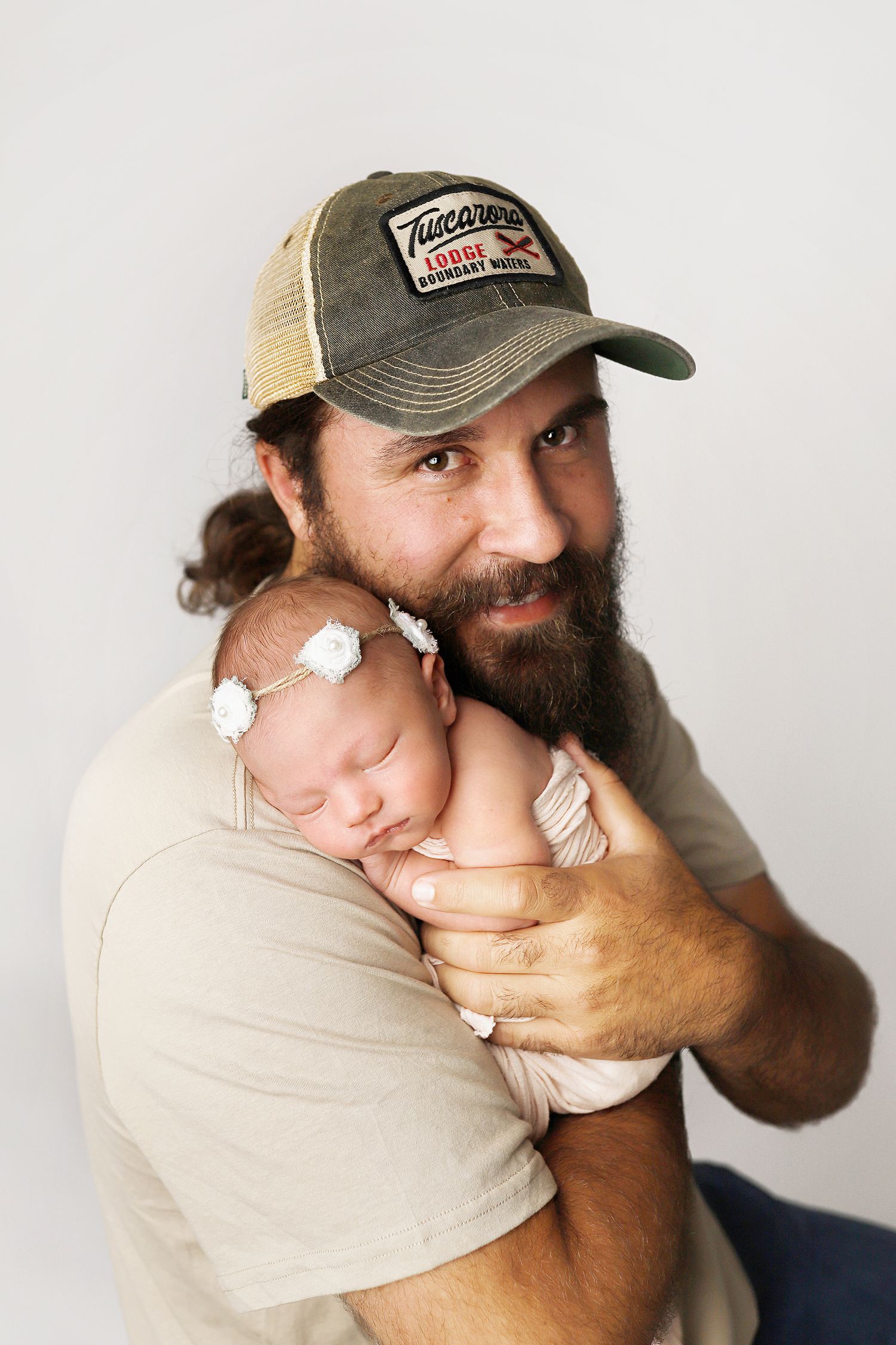 Tender portrait of a bearded person wearing a trucker hat holding a sleeping newborn baby wearing a white headband.