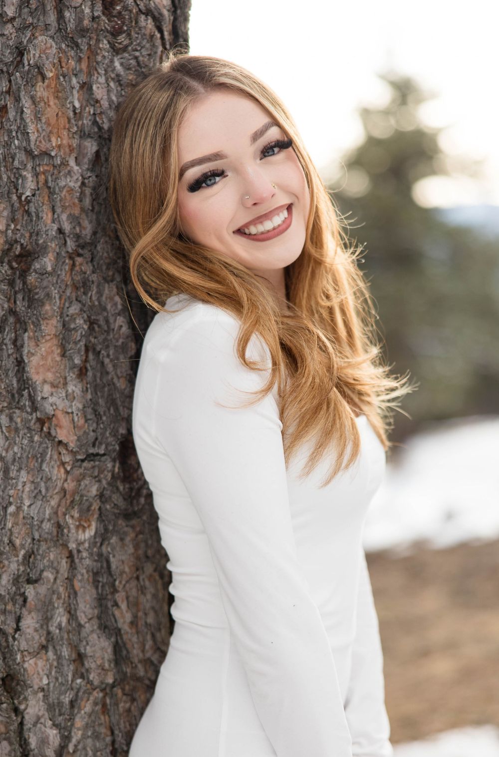 Portrait of woman in white sweater leaning against tree trunk with natural winter setting.