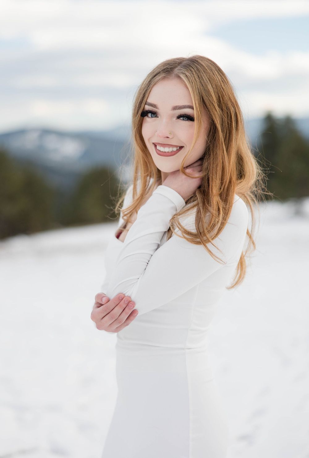 Woman in fitted white dress smiles brightly against winter mountain landscape.