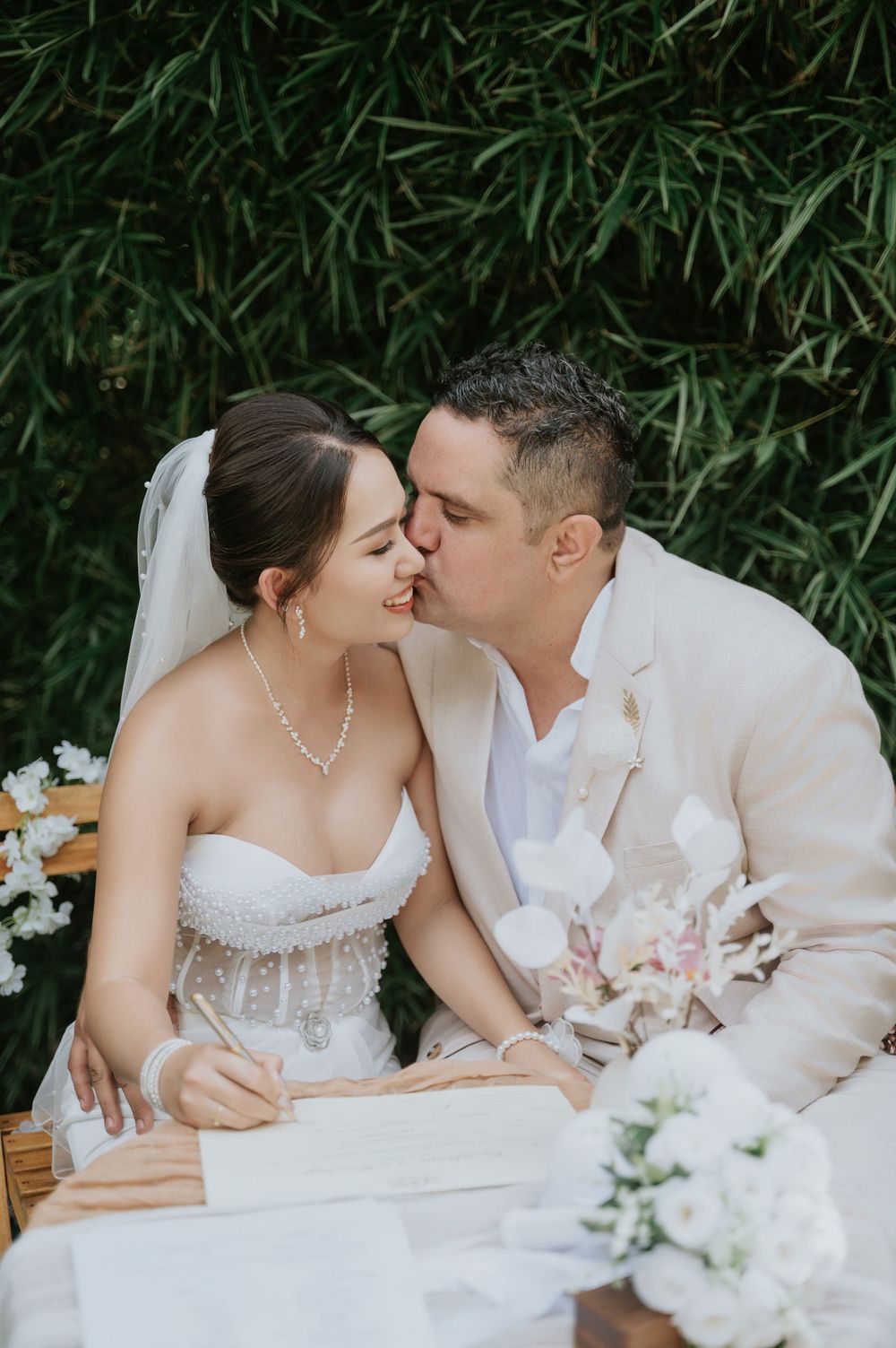 Newlyweds share an intimate kiss while signing their marriage certificate outdoors with bamboo backdrop and floral decor.