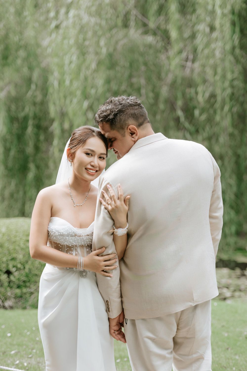 A tender moment between newlyweds in a garden setting with willow trees, both wearing elegant white and cream outfits.