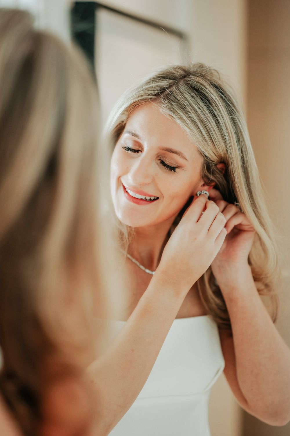Bride in white dress smiles with closed eyes while putting on earrings in soft warm lighting.
