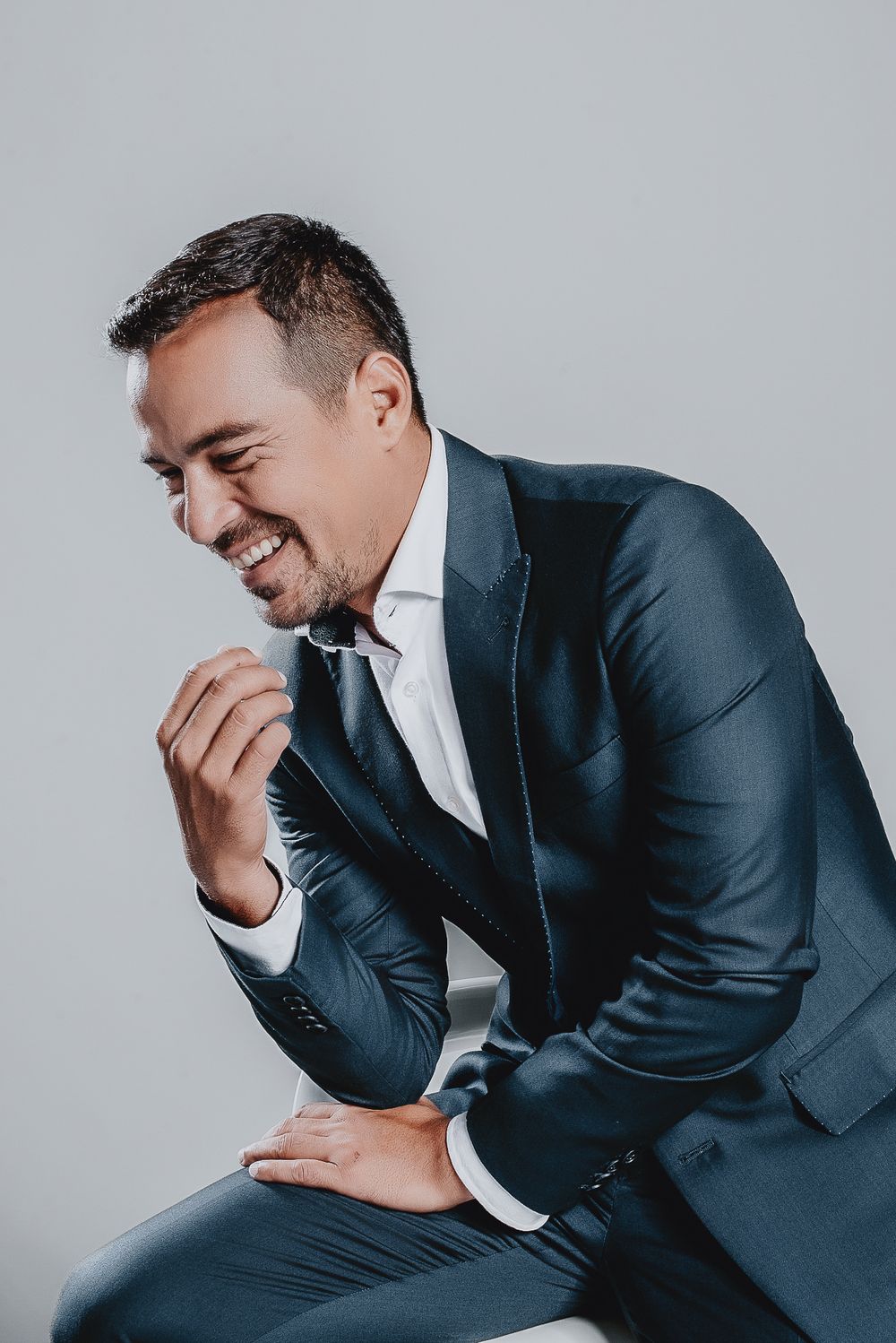 A person in a navy blue suit sits and laughs joyfully against a gray background in a professional portrait.