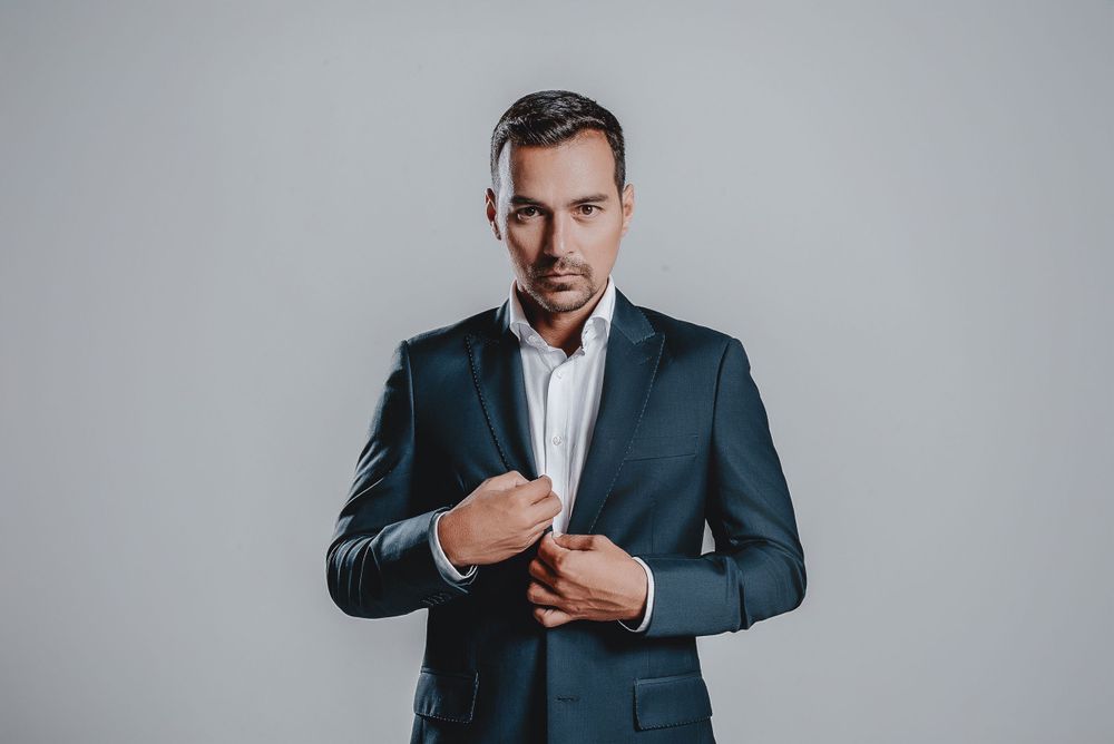 Professional in a dark suit adjusts his cuffs while standing against a gray backdrop.