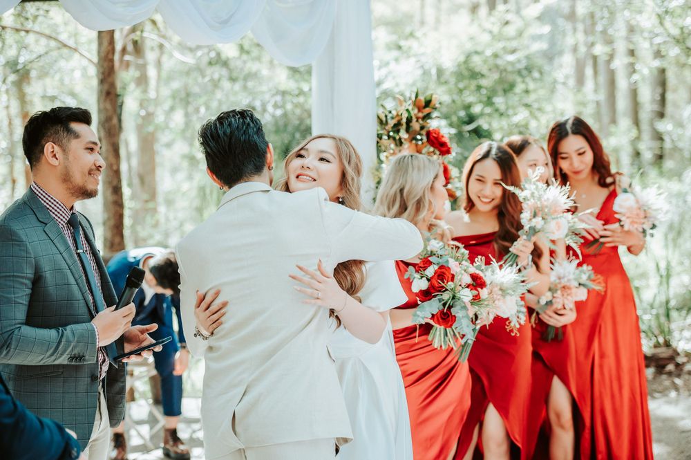 Wedding party celebrates joyfully outdoors with bridesmaids in vibrant red dresses holding bouquets against forest backdrop.