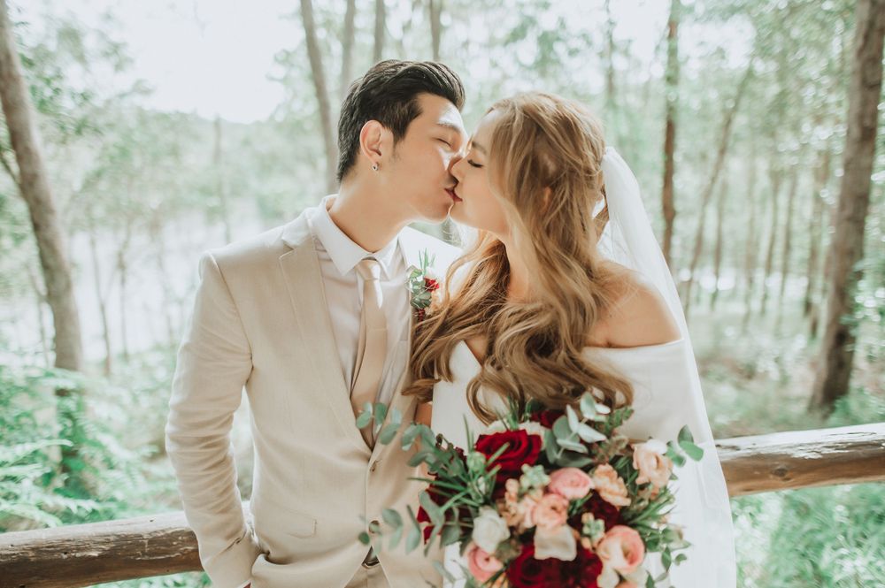 Romantic outdoor wedding portrait shows couple sharing intimate moment with floral bouquet in wooded setting.